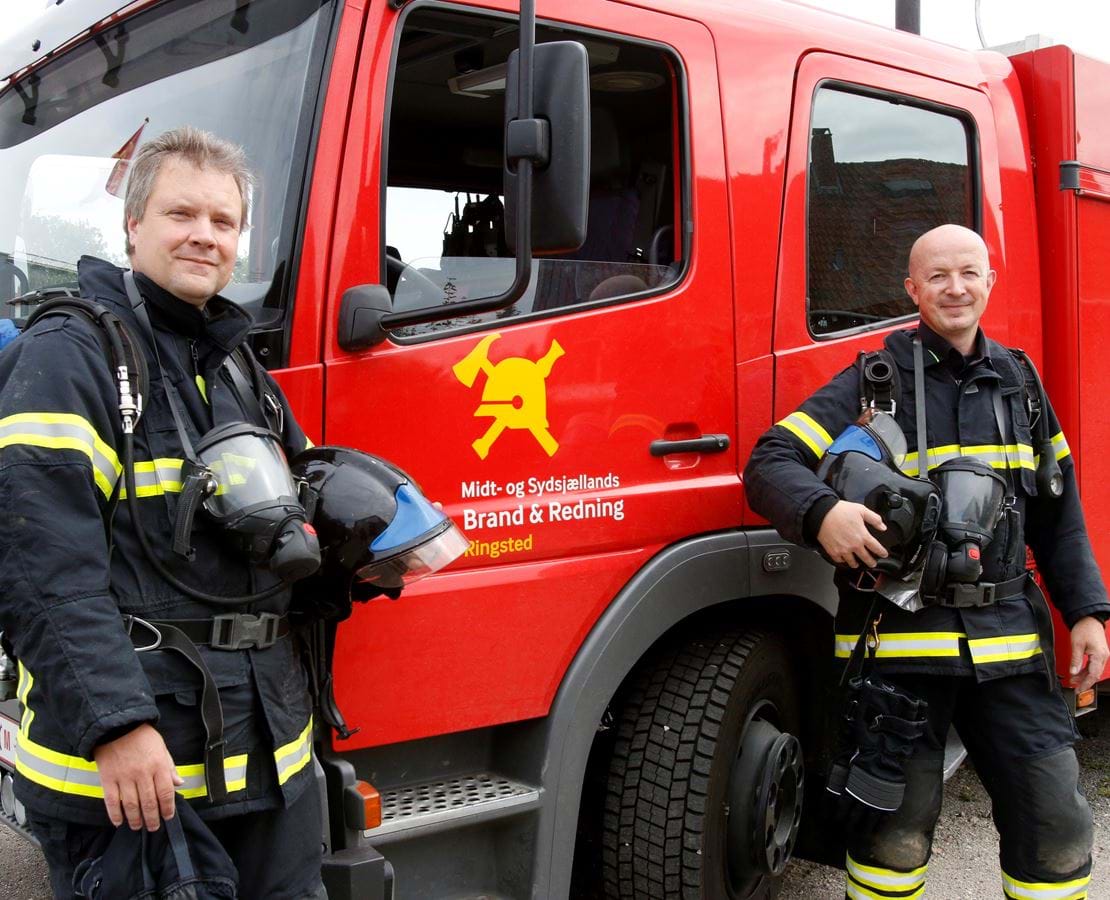To brandmænd står foran en rød brandbil og smiler.